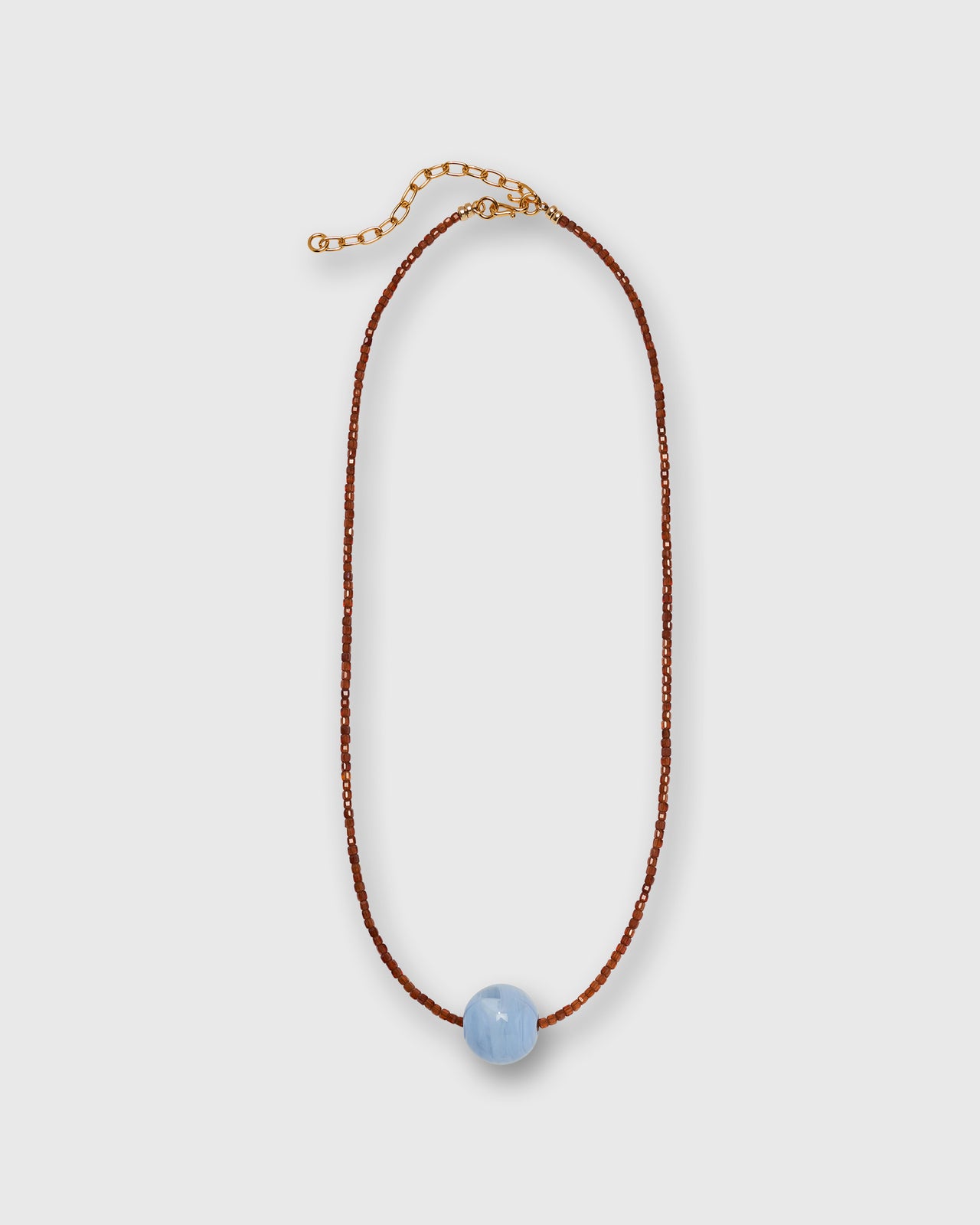 A delicate necklace featuring a thin reddish brown beaded cord, a gold-tone extender chain, and a single smooth, light blue bead at the center, displayed on a neutral light gray background.