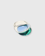 Load image into Gallery viewer, A sculptural, organic-shaped ring made of transparent smoky glass with a light blue center and a deep teal-green lower section, set against a solid light gray background.
